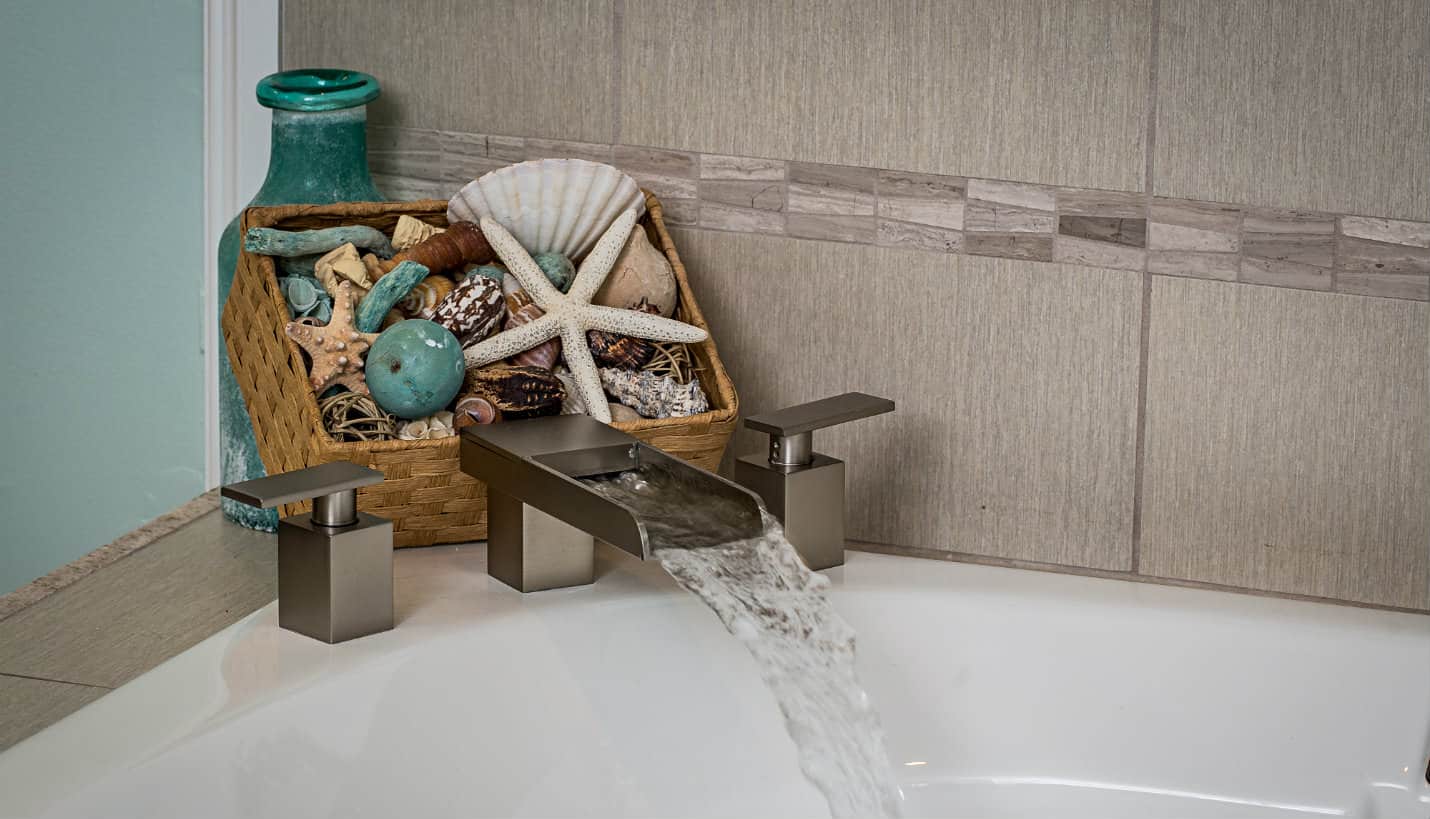 Silver rectangular faucet with flowing water into a tub and a basket of shells and starfish with an aqua bottle