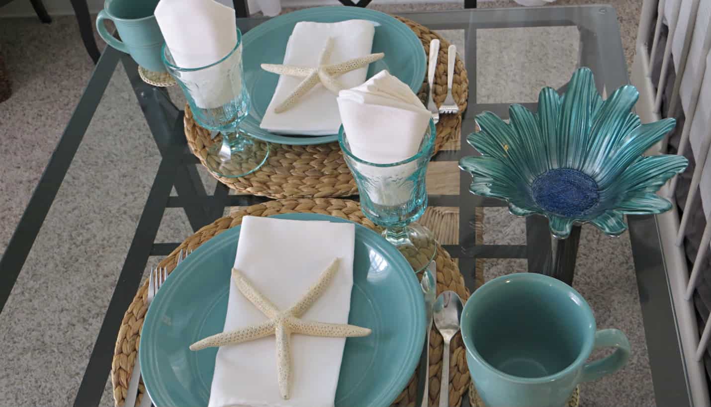 Black table with glass top, two aqua plates, cups and glasses, white napkins with a large white starfish.