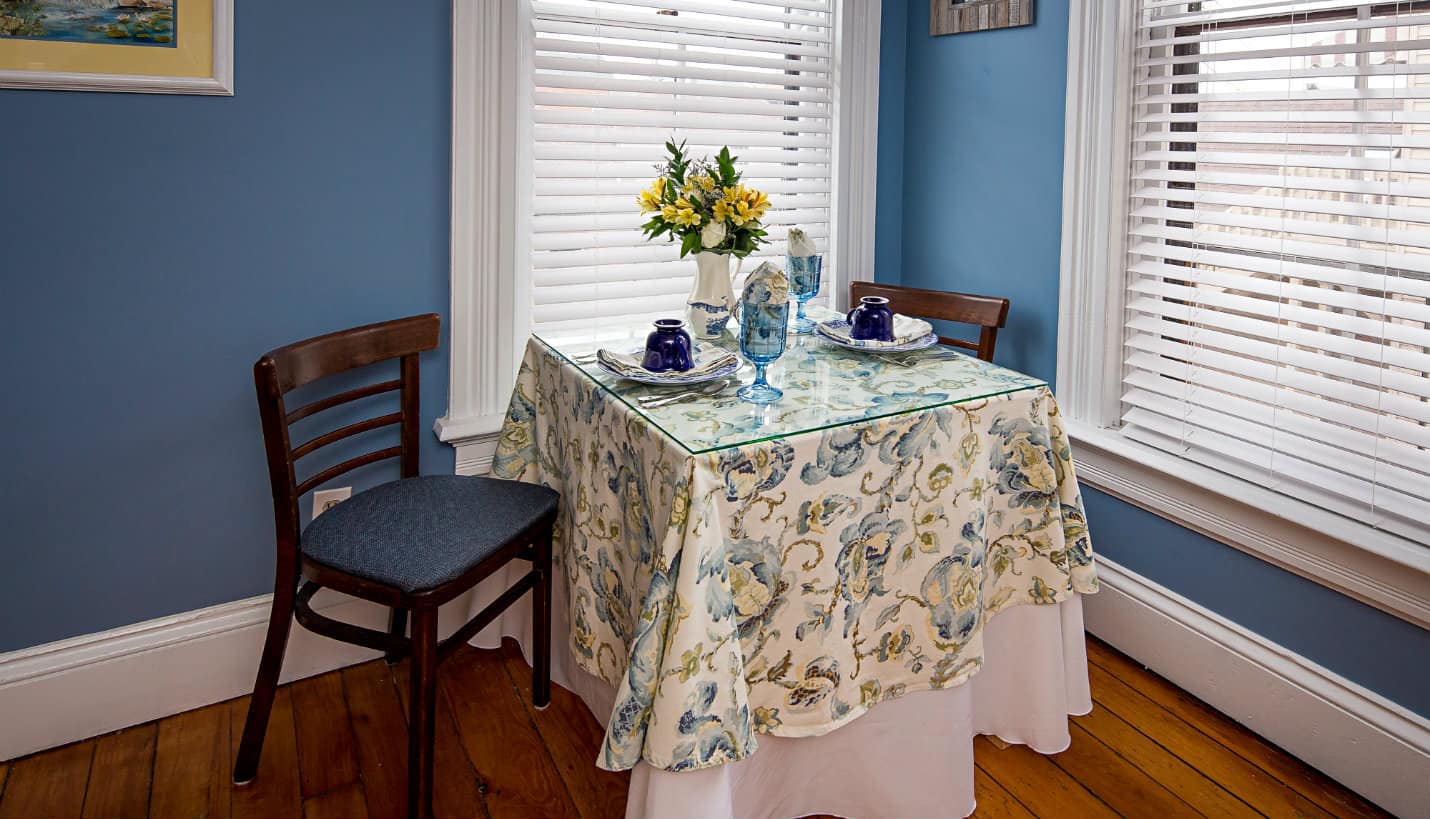 Table for two with blue and yellow tablecloth, between two tall white windows, two brown chairs with blue cushions