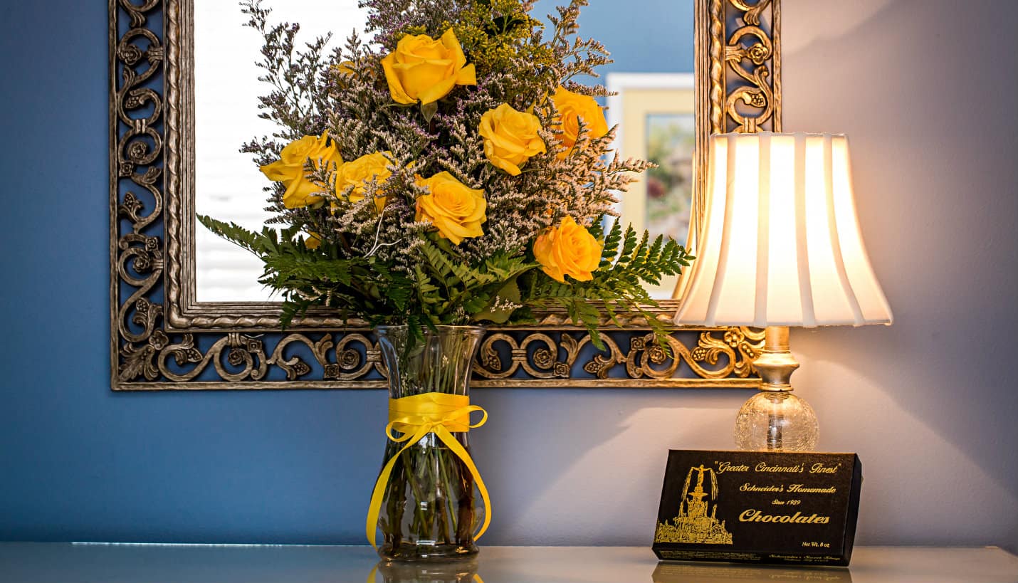 Large vase of yellow roses with green ferns in front of a silver scrolled mirror next to a lamp and box of candy