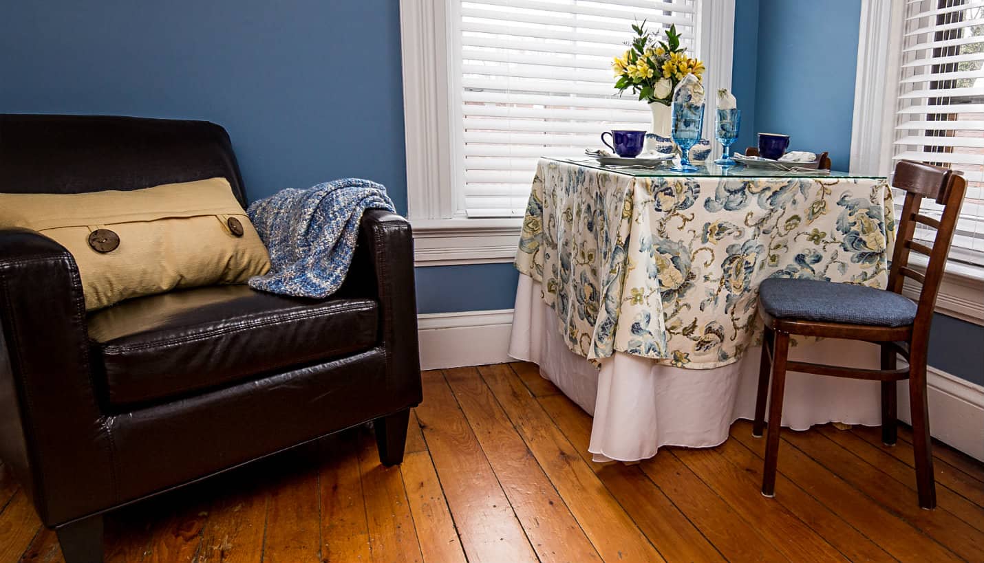 Brown chair with yellow pillow and blue quilt next to table for two with blue and yellow tablecloth