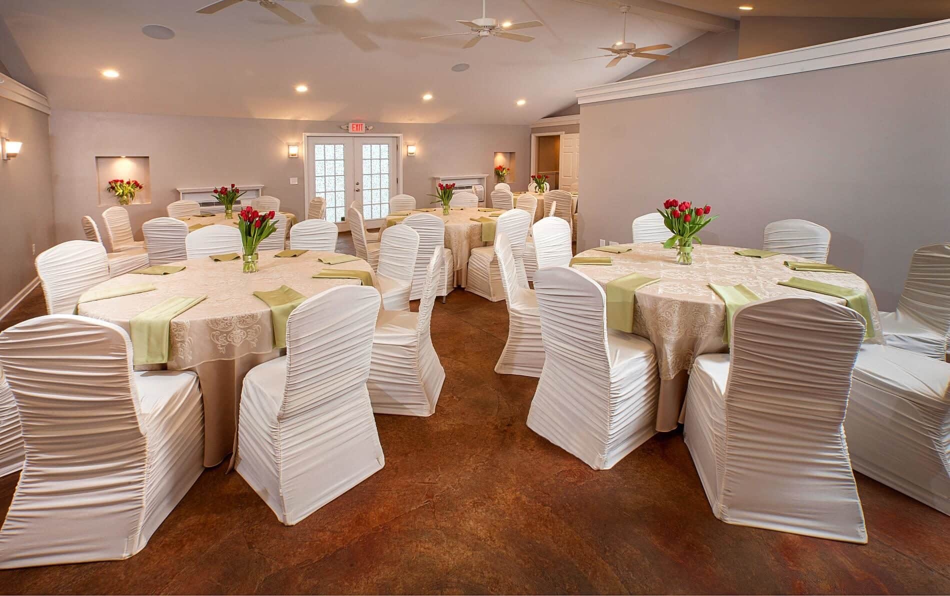 Large room with 6 round tables covered in beige lace tablecloths and white covered chairs