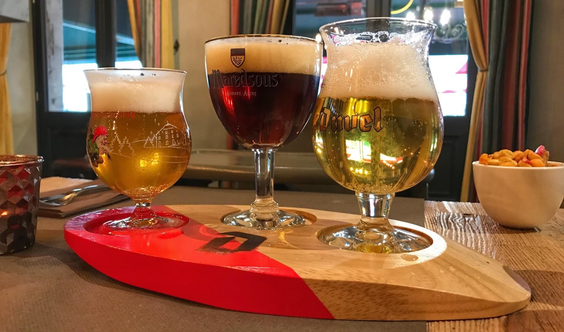 Three glasses of light and dark colored amber beer sitting on a red and tan wooden display tray