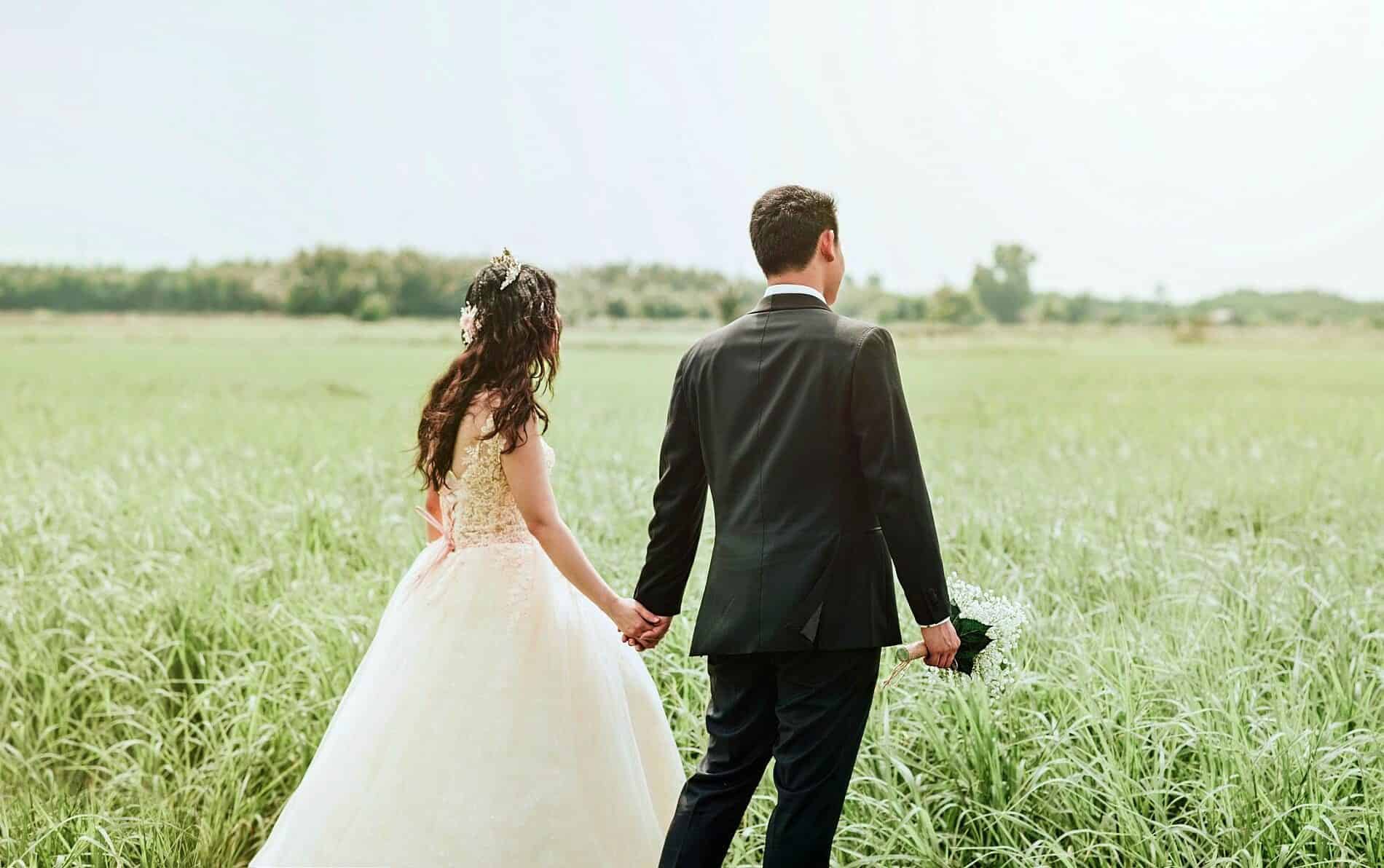 Bride in white dress holding hands with groom in black suite walking in open green field