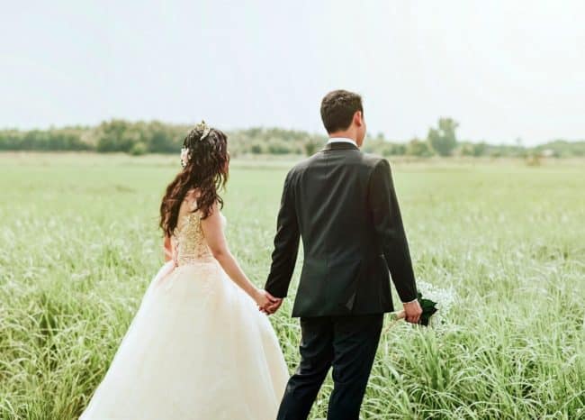 Bride in white dress holding hands with groom in black suite walking in open green field