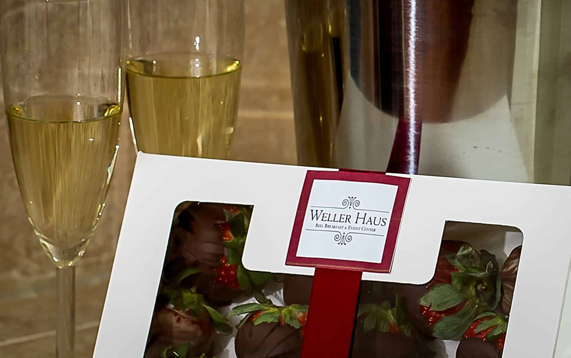 Two glasses of champagne with a white box tied with a red ribbon containing chocolate covered strawberries.