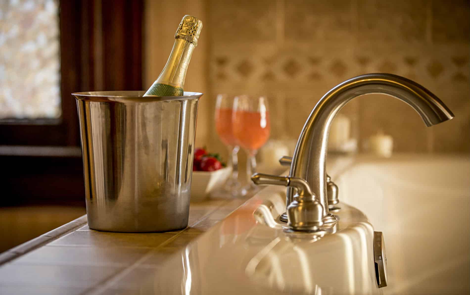 Large tub for two with a silver curved bathtub faucet. Silver bucket with champagne bottle , two glasses of pink champagne.