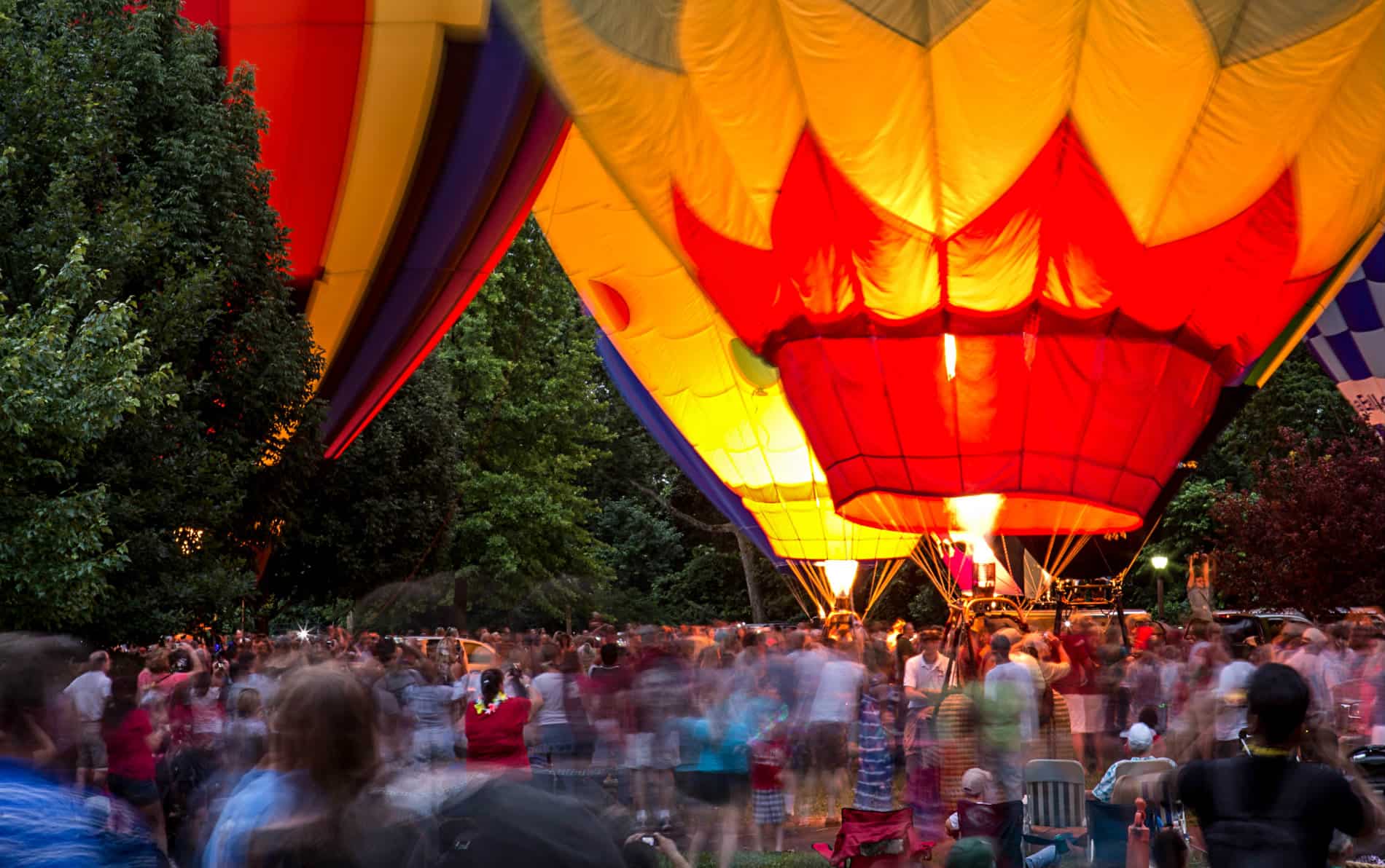 Red, yellow, green and blue hot air balloons surrounded by green trees and a crowd of people