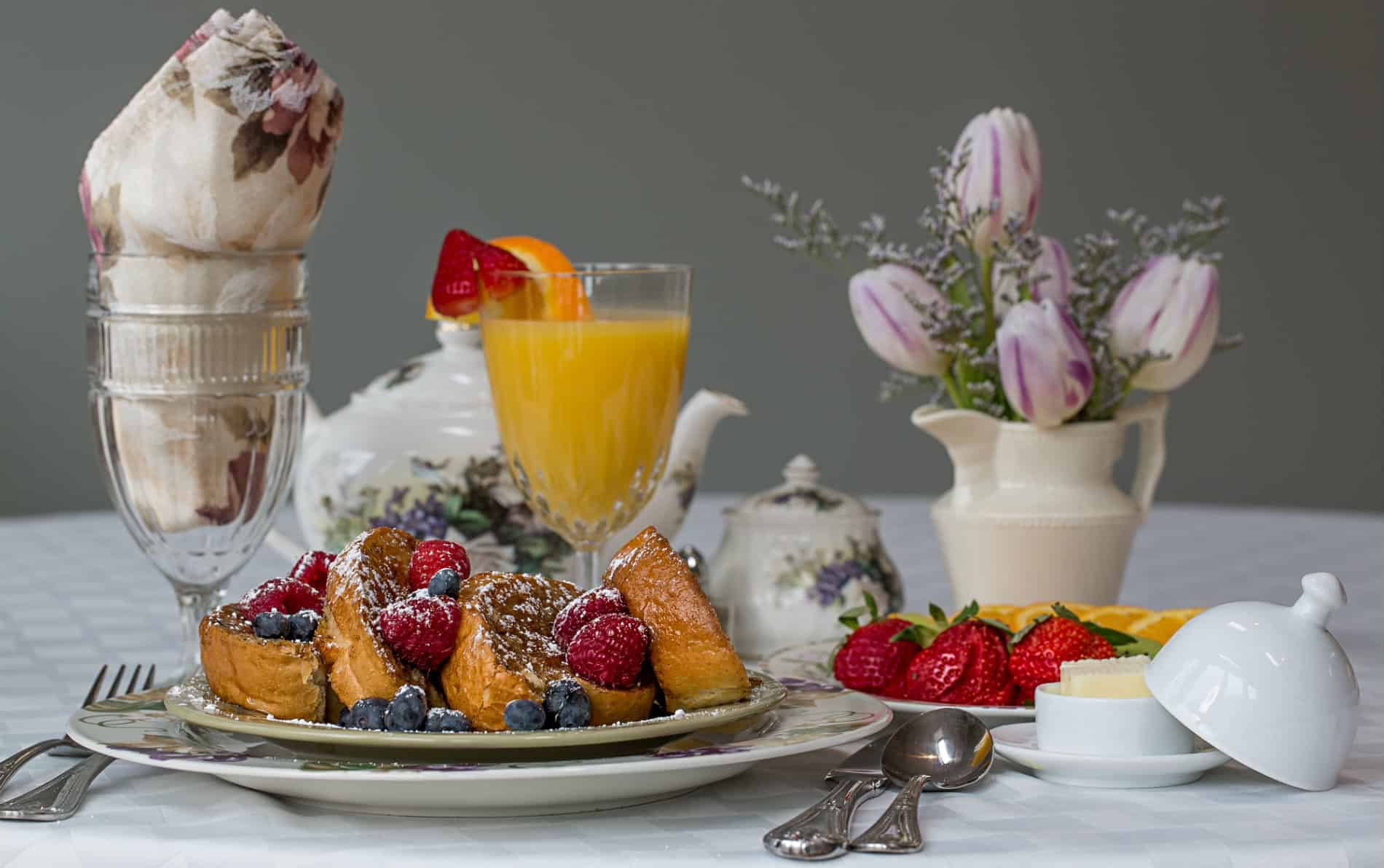 Sliced french toast with blueberries and raspberries, glass of orange juice, plate of sliced strawberries, small vase of purple tulips