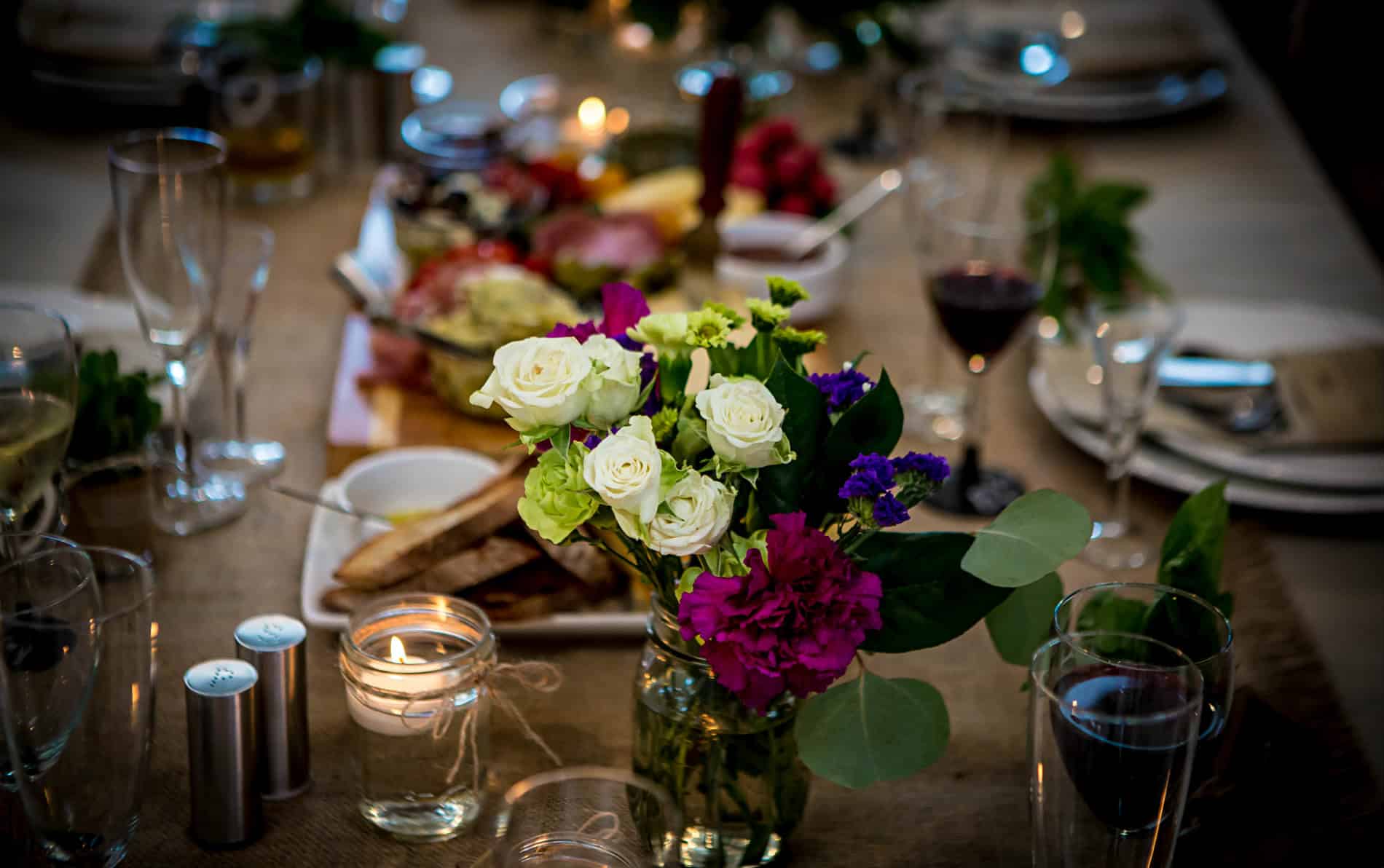 Dinner table set with red wine filled glasses, white plates, tan tablecloth, candles and white and purple flower arrangement.