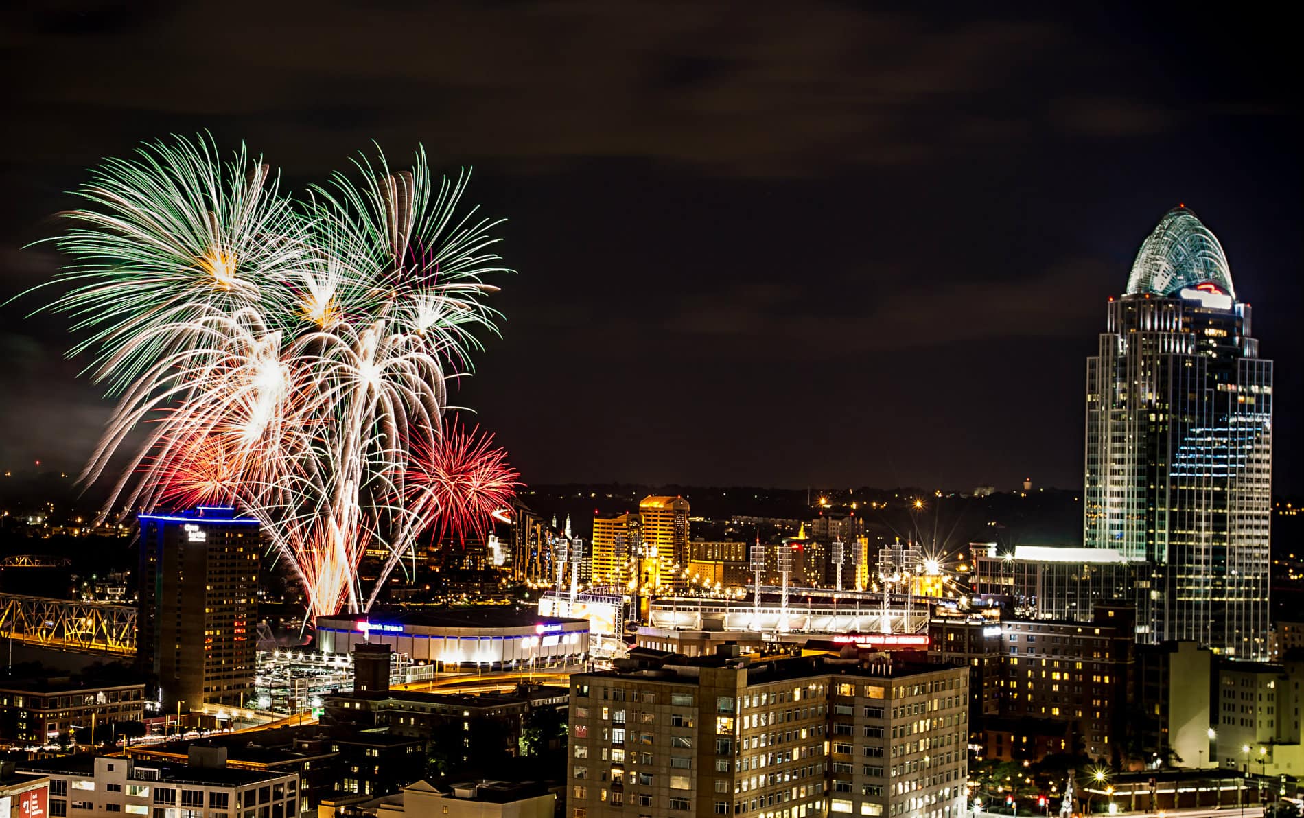 Lighted buildings with lights of blue, red, gold against a black night sky with red, green and white fireworks.