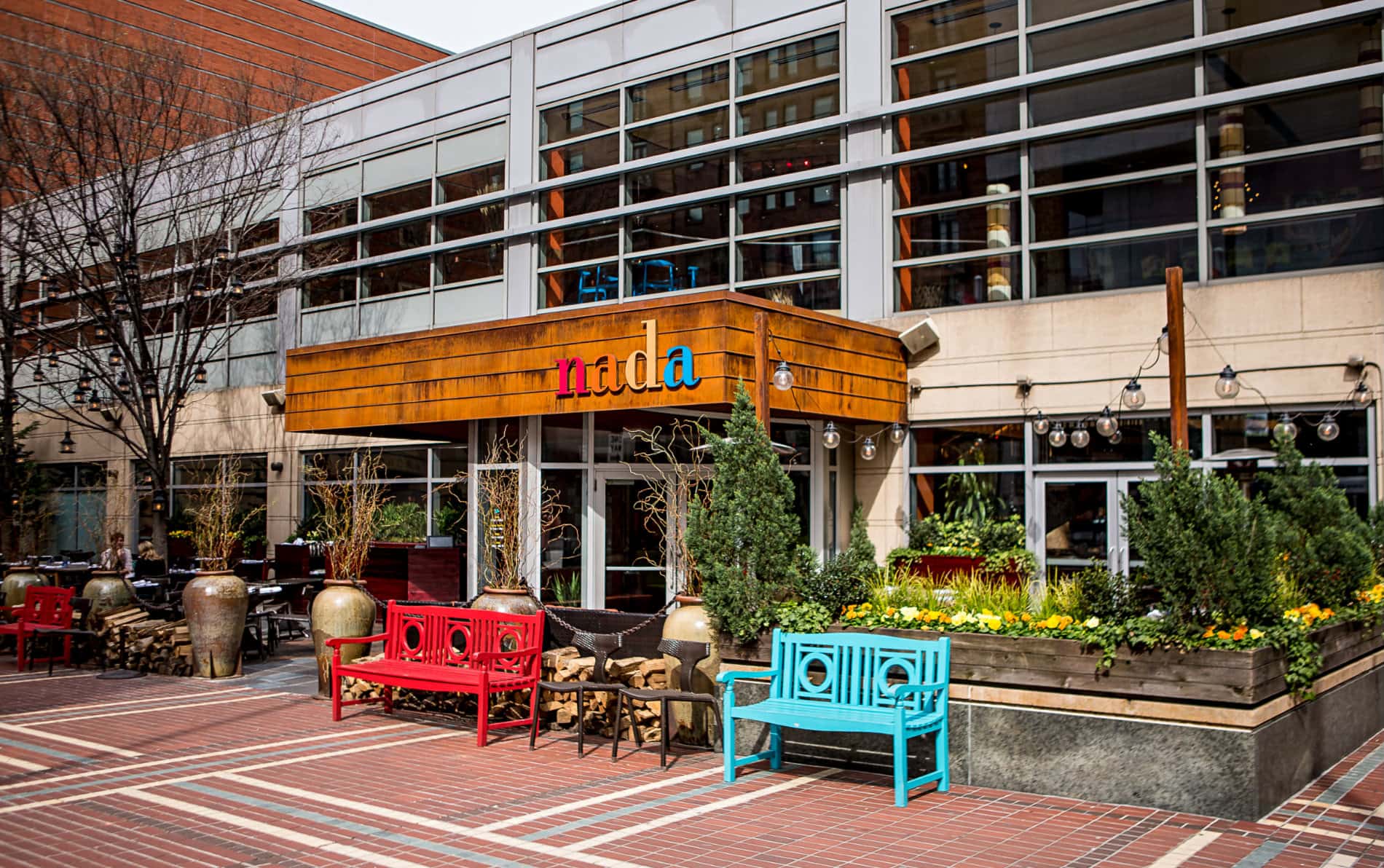 Long horizontal two-story restaurant building with 25 windows, aqua and red benches, planters with trees and yellow pansies