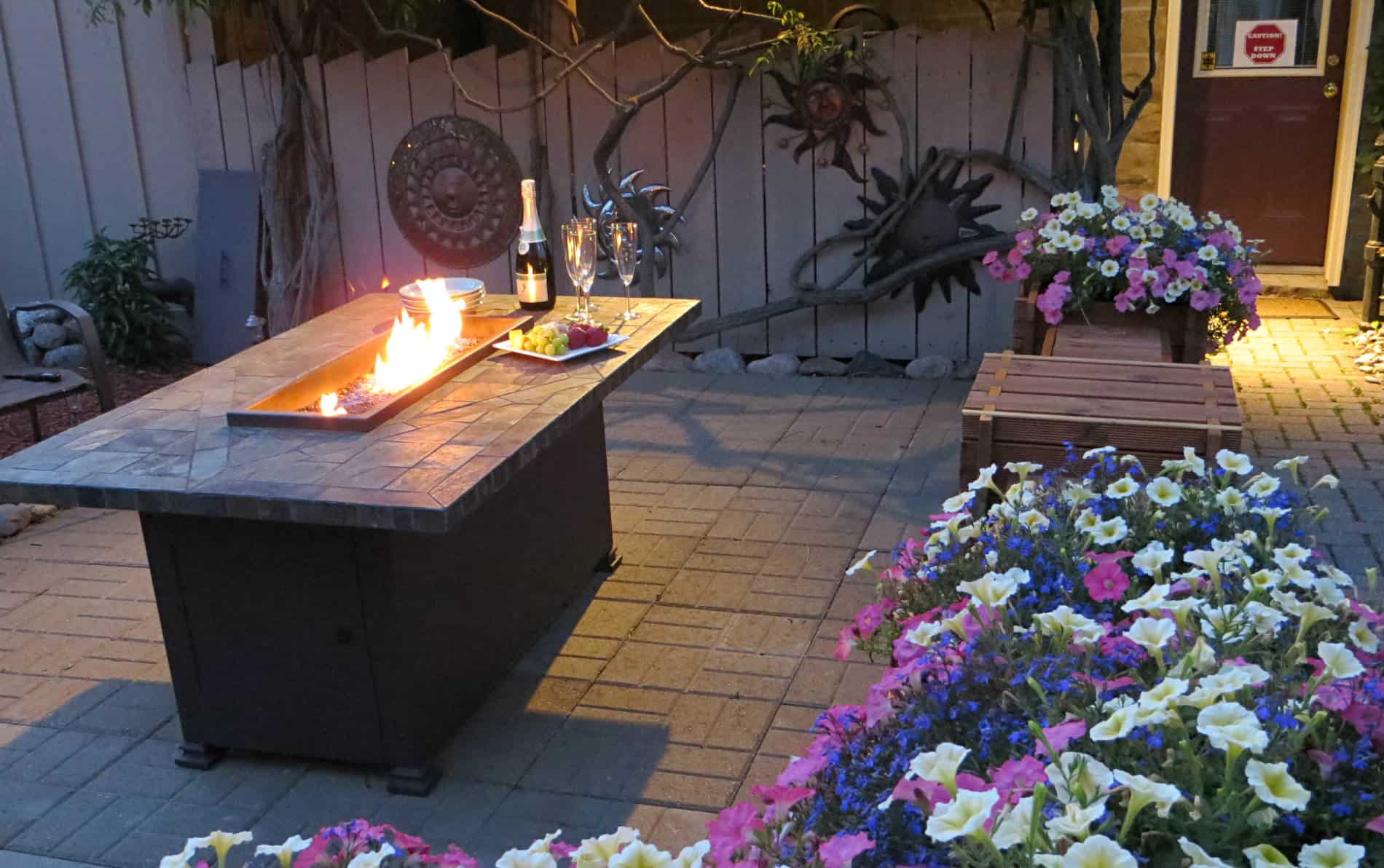 Rectangular table with fire in the middle, two glasses, champagne bottle and cheese tray with purple, pink and white flowers.