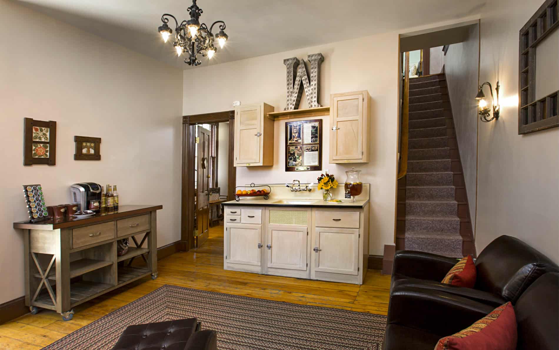 Small kitchen with three brown chairs with deep red pillows, multi colored braided rug, light colored cabinets, black iron chandelier