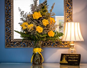 Large vase of yellow roses and a box of chocolates sitting in front of an ornate silver framed mirror.