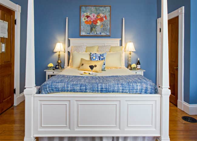 White four post bed with pale yellow and blue linens, against blue walls with a picture of yellow, pink, orange daisies