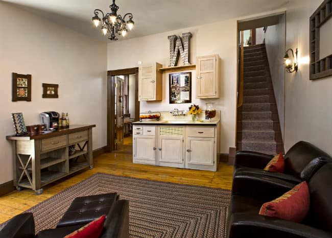 Small kitchen with three brown chairs with deep red pillows, multi colored braided rug, light colored cabinets, black iron chandelier