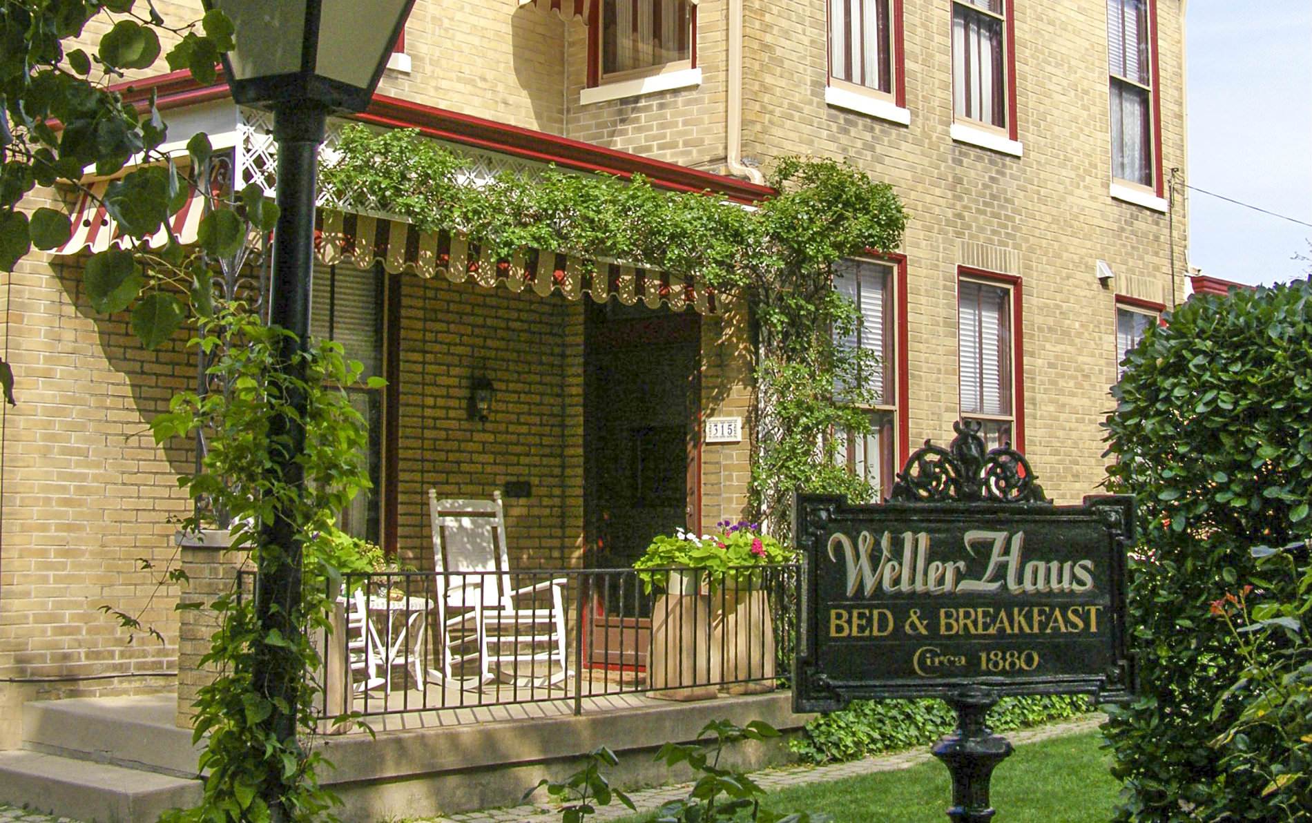 Two story tan brick house with six tall windows trimmed in red and white paint, porch with two rocking chairs