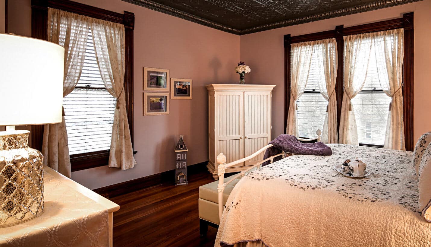 Large bedroom with hardwood floors, king-size off-white iron bed covered with off white quilt with pink embroidered roses