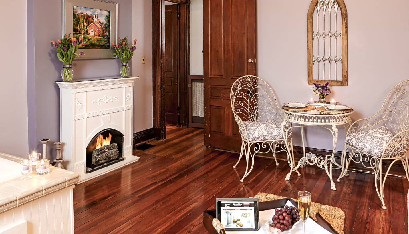 Large tub for two surrounded by lit candles, burning fireplace, and off white wrought iron table for two.