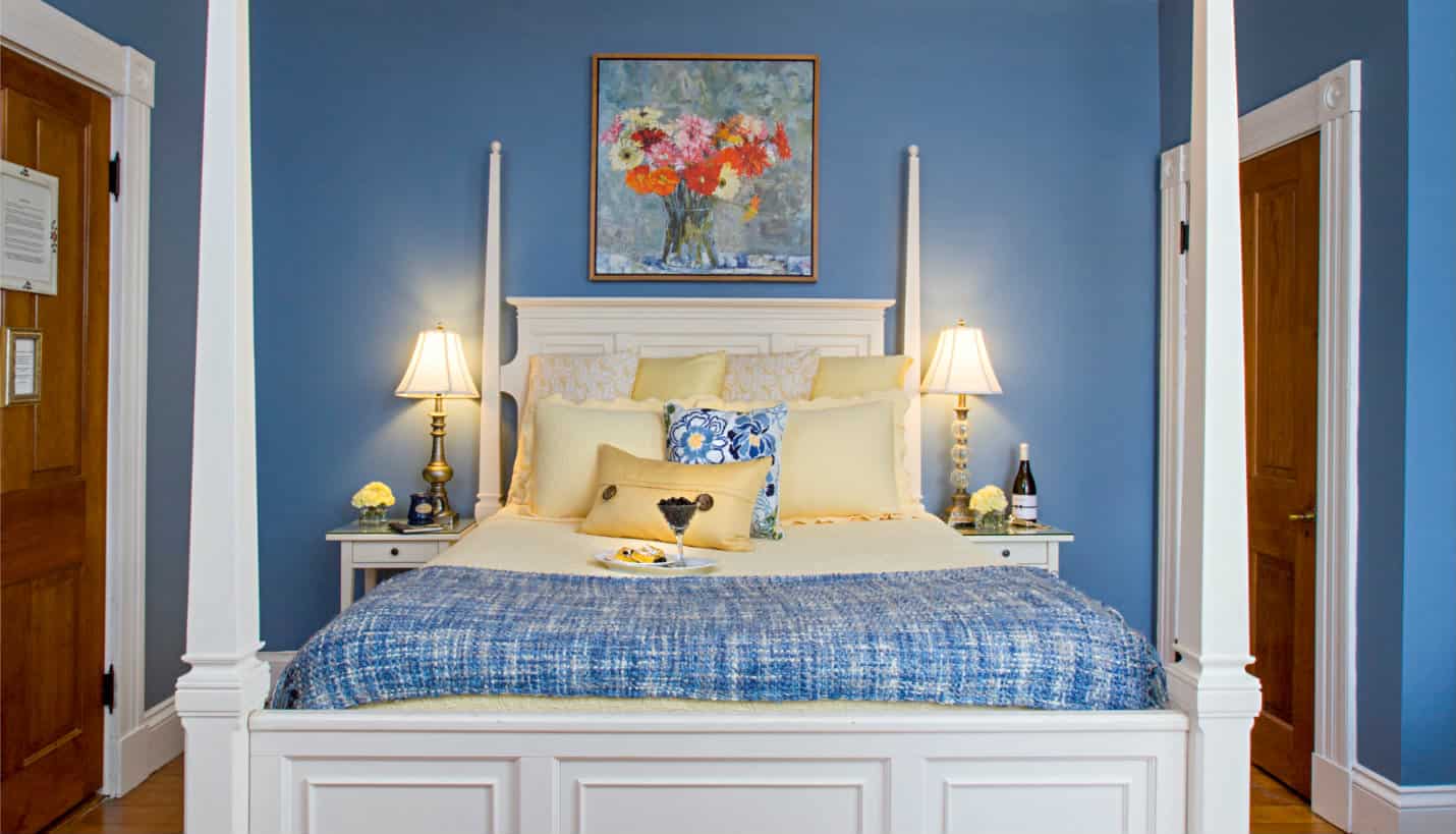White four post bed with pale yellow and blue linens, against blue walls with a picture of yellow, pink, orange daisies