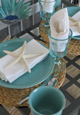 Breakfast table with two aqua place settings sitting on woven placemats, white napkins, decorative starfish and aqua drinking glasses.