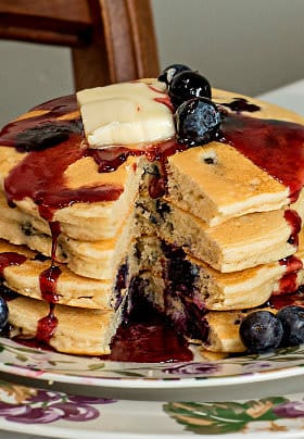 Small round plate with four stacked blueberry pancakes with blueberry sauce and a slice of yellow butter