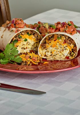 Scrambled eggs, sausage, cheese, red and green peppers inside a soft white tortilla topped with red tomatoes and cilantro.