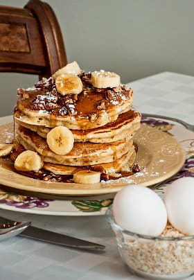 Four pancakes topped with pecans, sliced bananas, and syrup next to a cup of oatmeal grains topped with two eggs.