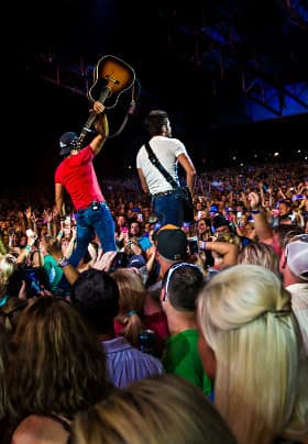 Two men with guitars, one playing and one holding the guitar in the air surrounded by a crowd of people