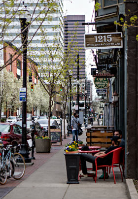 Street with trees in front of a restaurant. Sign with 1215 Wine Bar Coffee Lab, men sitting in red chairs