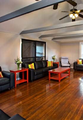 Room with hardwood floors, gray horizontal ceiling beams, black couches and red tables.