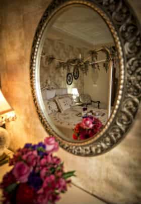 Oval silver mirror reflecting a white bed with an arrangement of pink and purple flowers