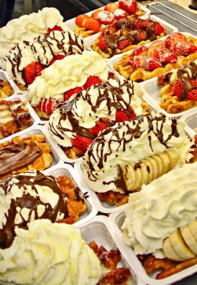 Fourteen white rectangular plates with thick waffles covered in bananas, strawberries, whip cream & chocolate