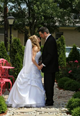 Bride in a white dress kissing her groom standing next to a pink table among evergreen trees and pink rosebushes