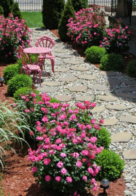 White rock and stone walkway surrounded by pink rosebushes on each side and a pink table & chairs on walkway