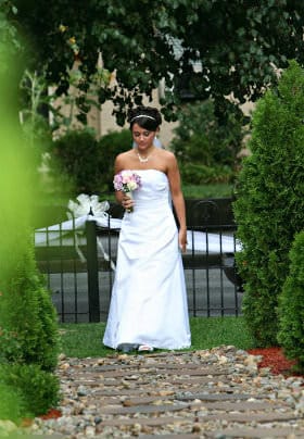 Bride in a white dress with purple flowers walking down a stone bathway surrounded by tall evergreen bushes