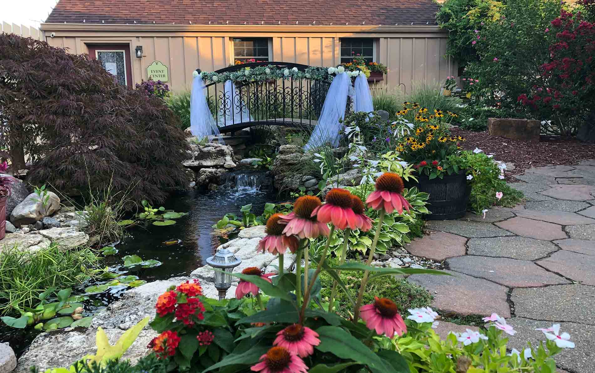 Garden view over stream with pathway lined with colorful blossoms leading to bridge over stream. 